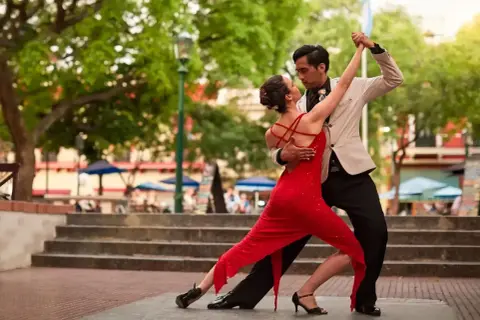 Para tańczy tango na zewnątrz, kobieta w czerwonej sukience, mężczyzna w jasnej marynarce.