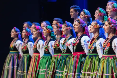 Zespół ludowy w tradycyjnych strojach, kobiety w kolorowych chustach i spódnicach, mężczyźni w tle.