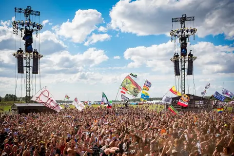 Tłum ludzi na festiwalu pod sceną z wieżami nagłośnieniowymi i flagami.