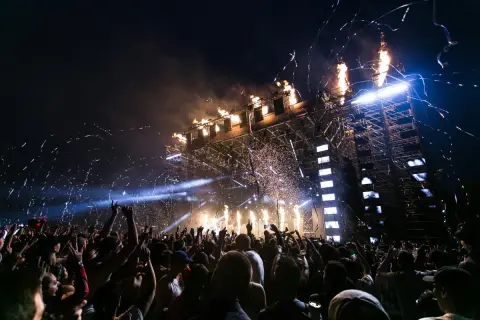 Tłum ludzi na koncercie nocą, z fajerwerkami i konfetti nad sceną.