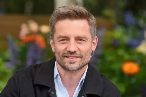 Marcin Rogacewicz, wiek nieznany, uśmiecha się do kamery w otoczeniu zieleni i kolorowych kwiatów.