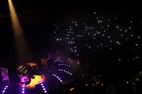 Koncert w ciemności: scena z muzykami, publiczność z zapalonymi telefonami.