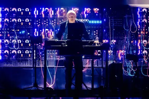 Hans Zimmer stoi przy syntezatorze na tle ściany pełnej migających świateł i kabli.