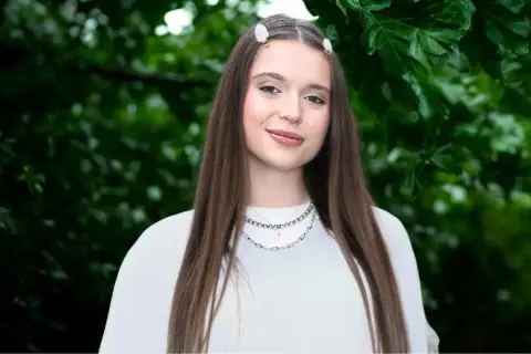 Uśmiechnięta Nicole Pniewska w białej bluzce, z długimi brązowymi włosami i delikatnymi spinkami, na tle zielonych liści.