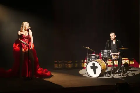 Kasia Gołomska w czerwonej sukni śpiewa przy mikrofonie, a za nią perkusista gra na bębnach.