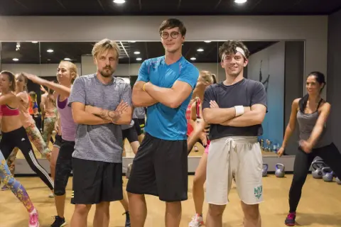 Trzech mężczyzn w strojach sportowych stoi z rękami skrzyżowanymi na piersi, w tle grupa kobiet ćwiczy.
