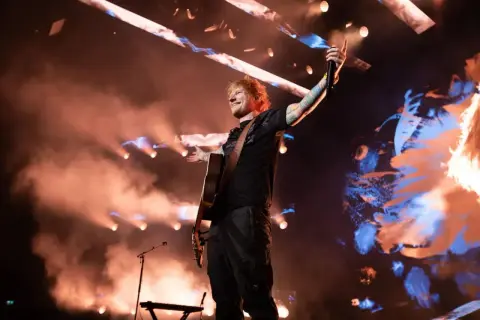 Ed Sheeran na koncercie w Polsce, grając na gitarze i pozdrawiając publiczność.