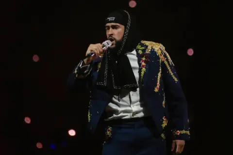Bad Bunny w ozdobnej marynarce śpiewa na koncercie na ciemnym tle z rozmazanymi światłami.