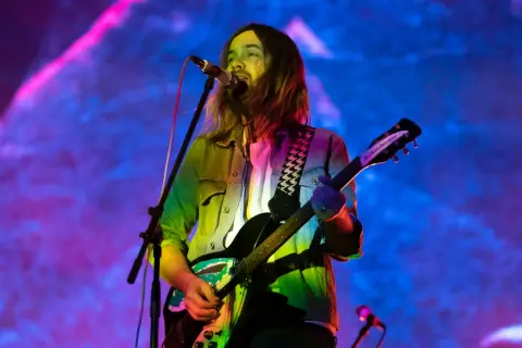 Kevin Parker z Tame Impala gra na gitarze podczas koncertu, w tle kolorowe światła.