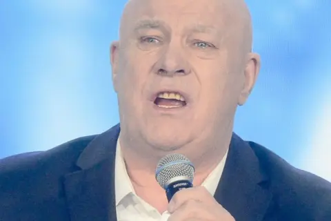Ryszard Rynkowski śpiewa do mikrofonu, ubrany w marynarkę i białą koszulę.