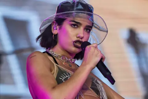 Dua Lipa w przezroczystym kapeluszu śpiewa do mikrofonu na koncercie.