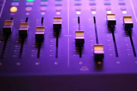 Panel miksera dźwiękowego z faderami i podświetlanymi przyciskami w fioletowym świetle.