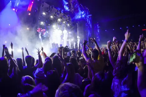 Tłum ludzi z rękami w górze na koncercie, scena z efektami świetlnymi i dymem.