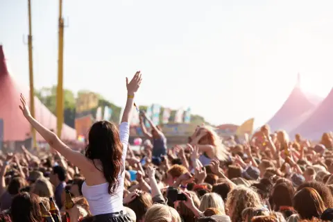 Tłum ludzi na festiwalu muzycznym, z rękami w górze, w tle namioty i sceny.
