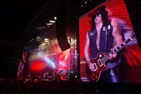Slash z Guns N' Roses gra na gitarze na wielkim ekranie podczas koncertu.