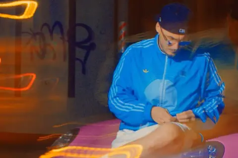 Mężczyzna w niebieskiej kurtce i czapce siedzi na tle nocnego miasta z neonowymi światłami.