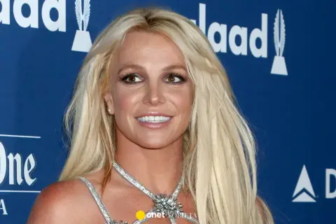 Uśmiechnięta Britney Spears, której wiek nie jest widoczny na zdjęciu, w błyszczącej kreacji na tle niebieskiej ścianki z logo GLAAD.
