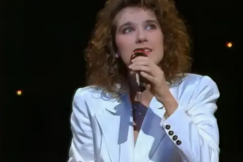 Celine Dion w białej marynarce śpiewa do mikrofonu na ciemnej scenie.