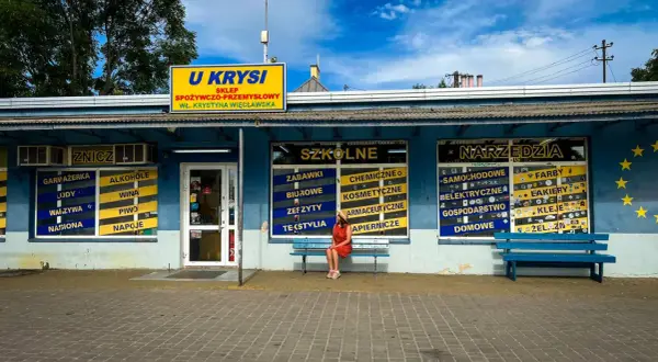 Dziewczyna w czerwonej sukience siedzi na ławce przed sklepem "U Krysia", gdzie kręcono "Ranczo".