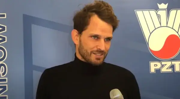 Maciej Synówka, z brodą i w czarnym golfie, udziela wywiadu na tle niebieskiej ściany z logo PZT.