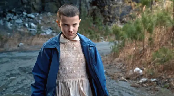 Nastka z Stranger Things, z krótkimi włosami i w niebieskiej kurtce, stoi na brudnej drodze.