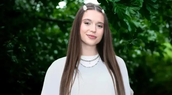 Uśmiechnięta Nicole Pniewska w białej bluzce, z długimi brązowymi włosami i delikatnymi spinkami, na tle zielonych liści.
