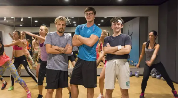 Trzech mężczyzn w strojach sportowych stoi z rękami skrzyżowanymi na piersi, w tle grupa kobiet ćwiczy.