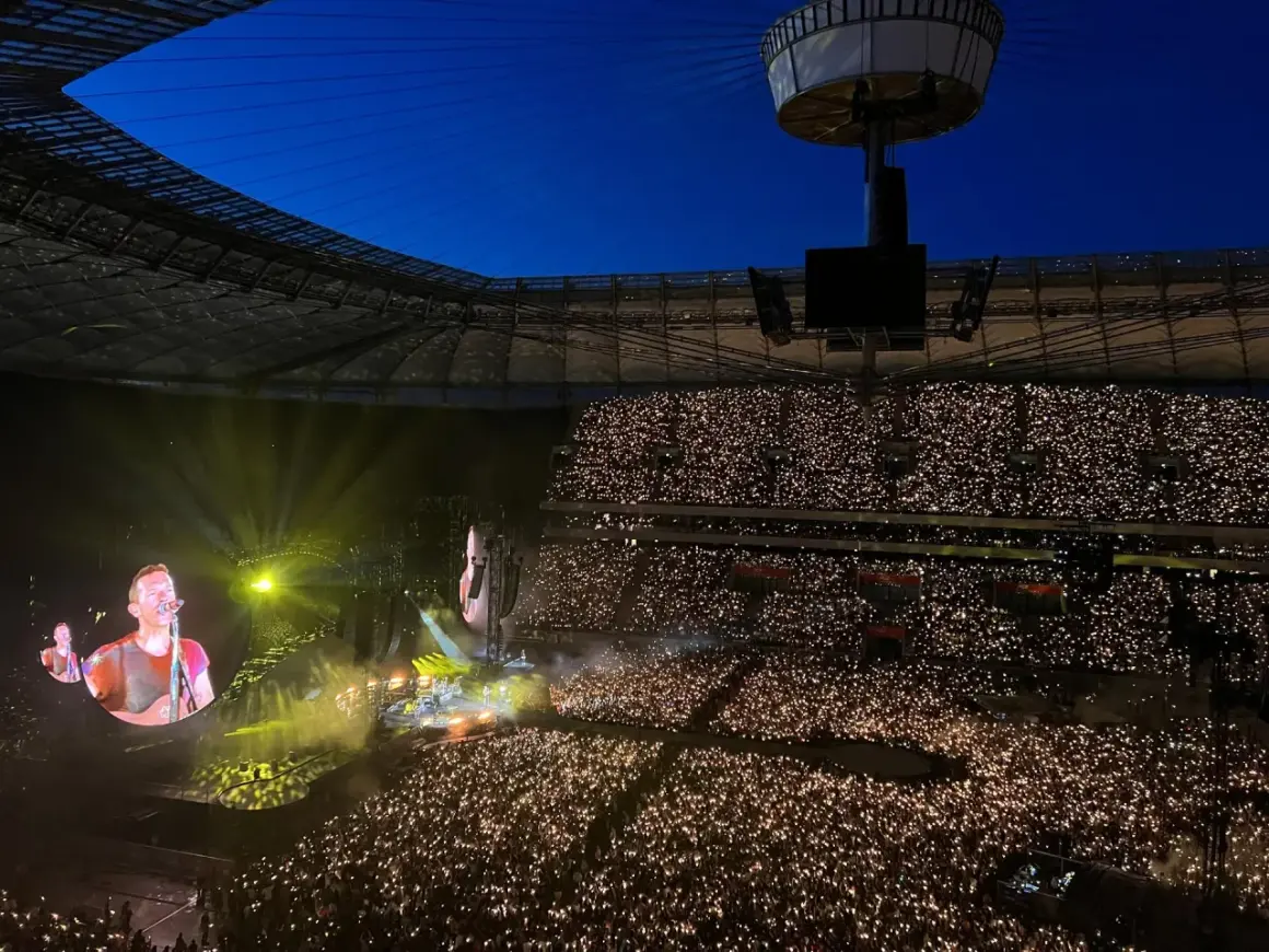 Koncert na stadionie, tłum z zapalonymi telefonami, na ekranie wokalista Coldplay.