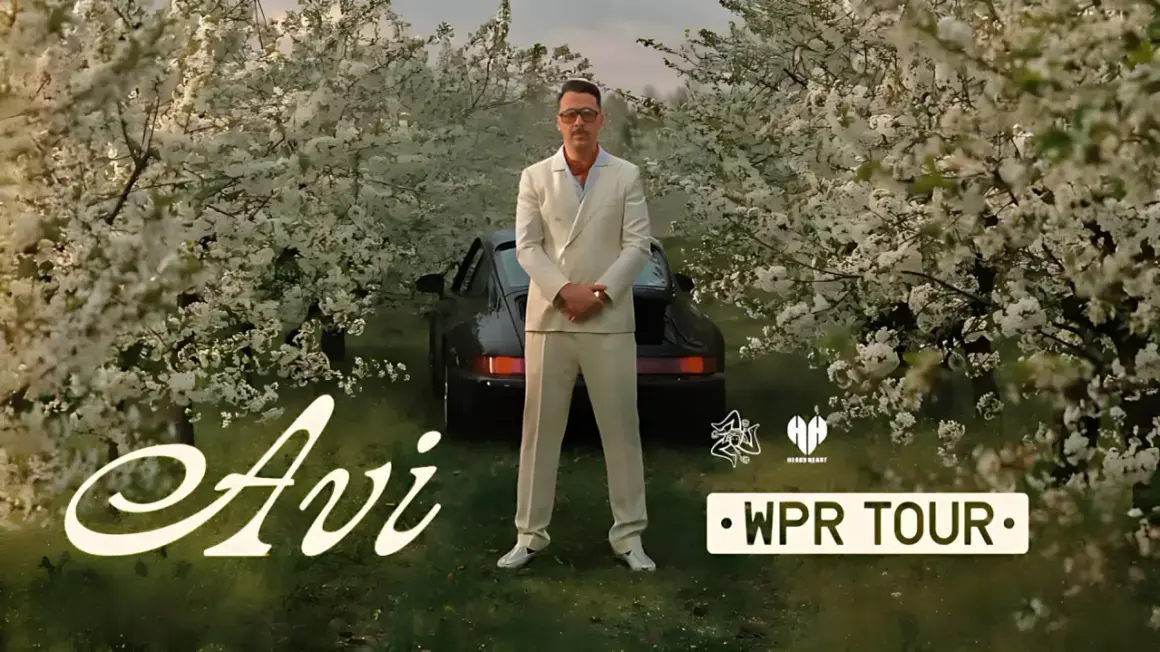 Mężczyzna w garniturze stoi przed czarnym samochodem wśród kwitnących drzew. Napis "Avi" i "WPR TOUR".