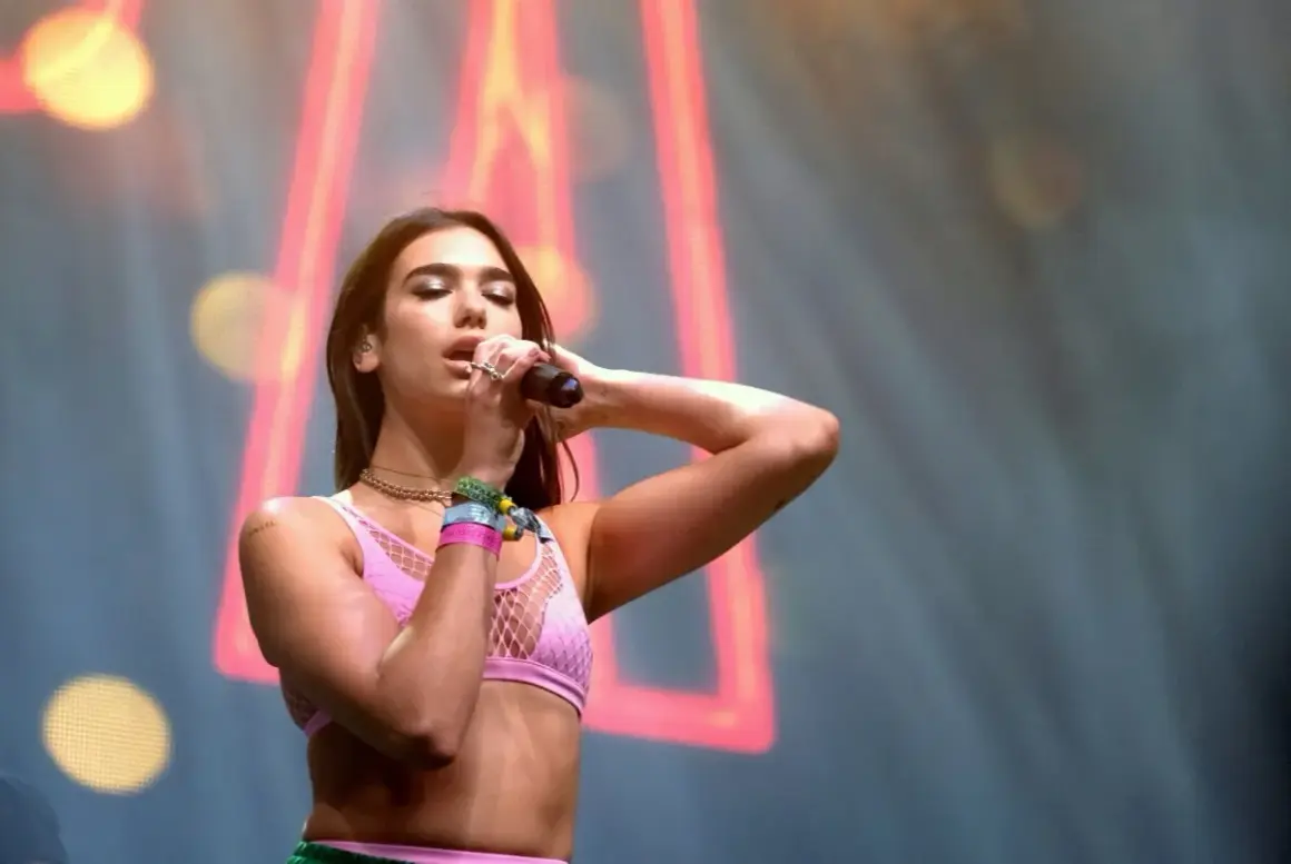Dua Lipa śpiewa na koncercie w różowym topie z siateczką, trzymając mikrofon.