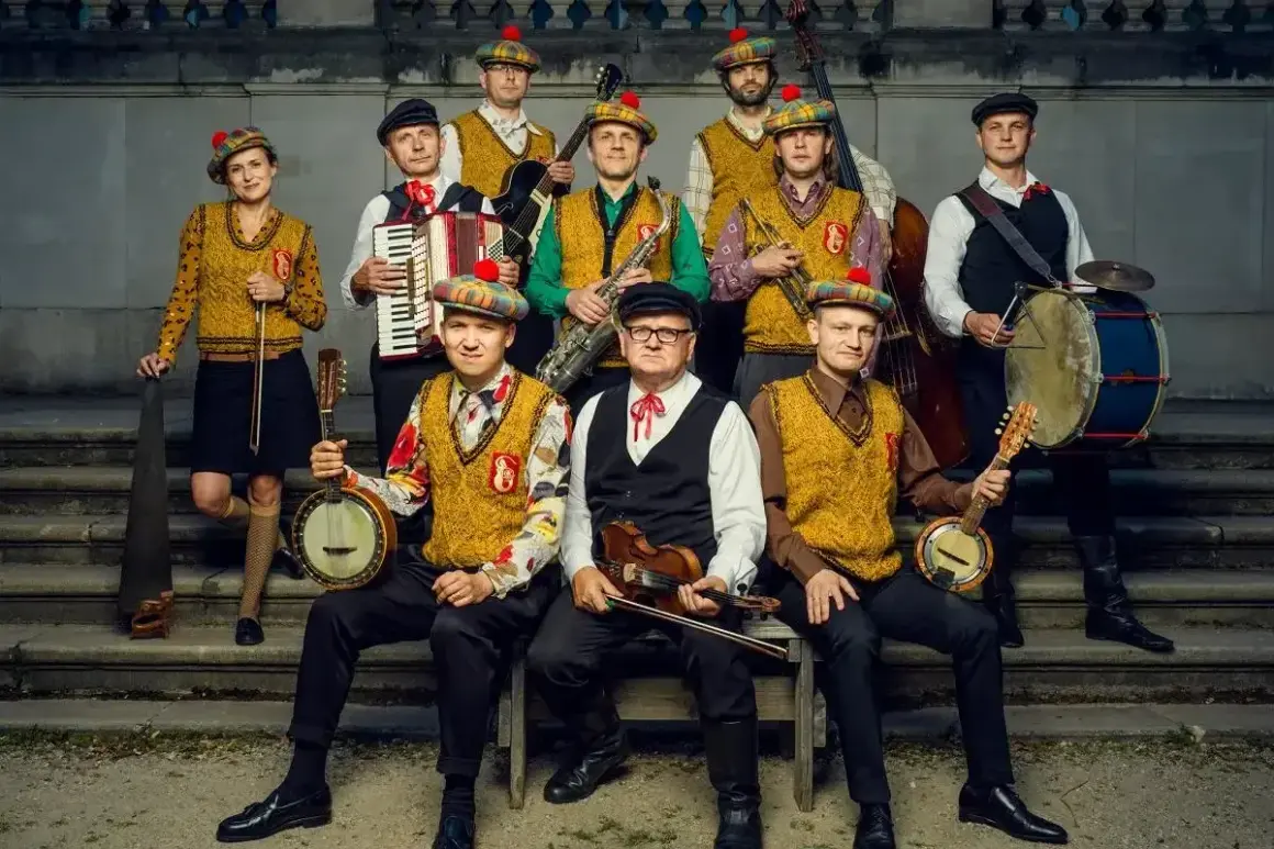 Zespół muzyczny w strojach ludowych z kapeluszami w kratę, grający na instrumentach: banjo, skrzypcach, akordeonie, saksofonie, kontrabasie i bębnie.
