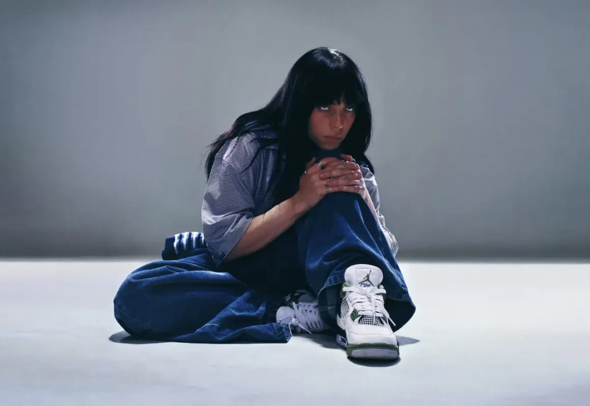 Billie Eilish siedzi na podłodze w luźnych dżinsach i koszuli, patrząc w obiektyw.