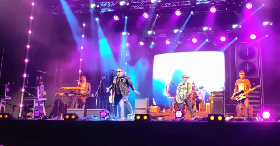 Zespół gra na scenie podczas koncertów w Stalowej Woli. Wokalista w skórzanej kurtce, gitarzyści i klawiszowiec.