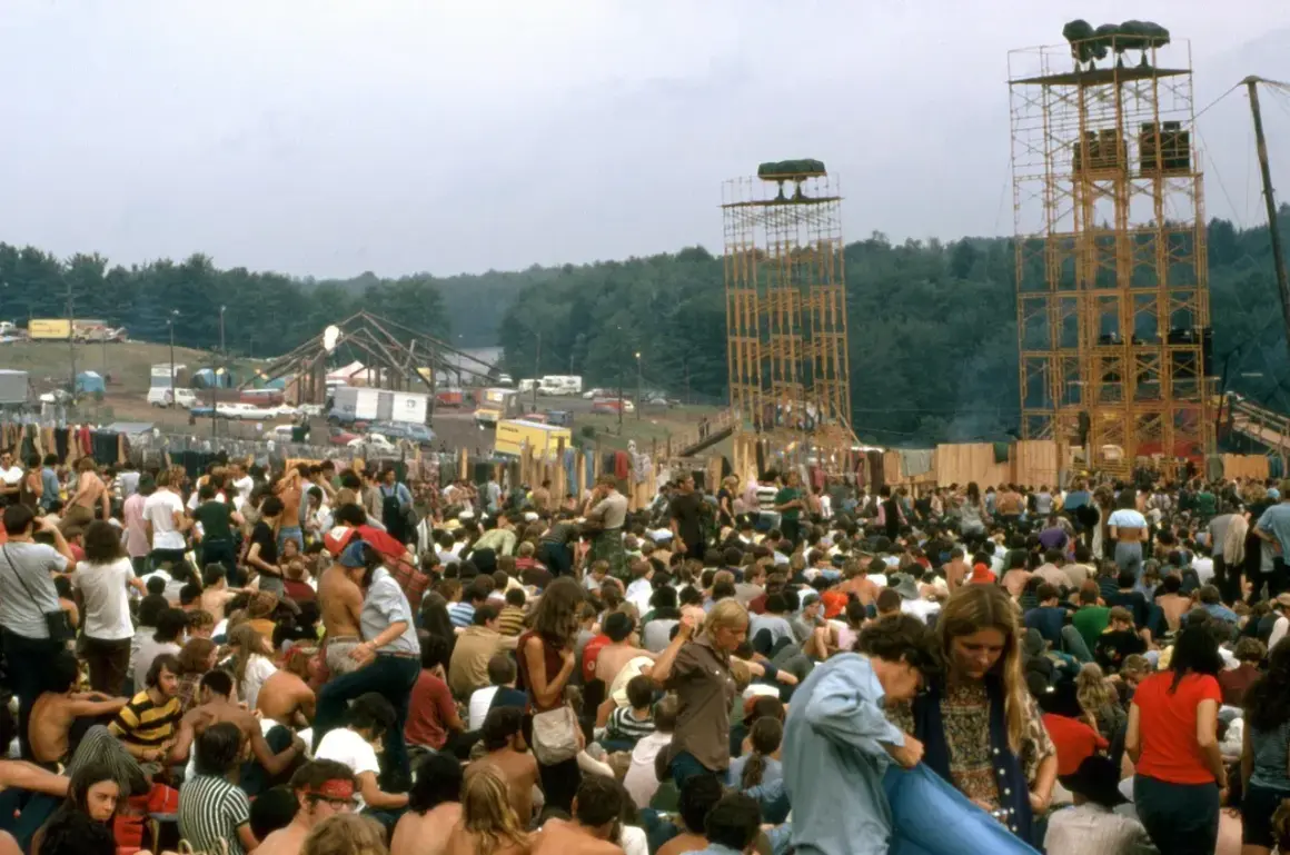 Tłum ludzi na festiwalu, widoczne wieże z głośnikami i scena w tle.