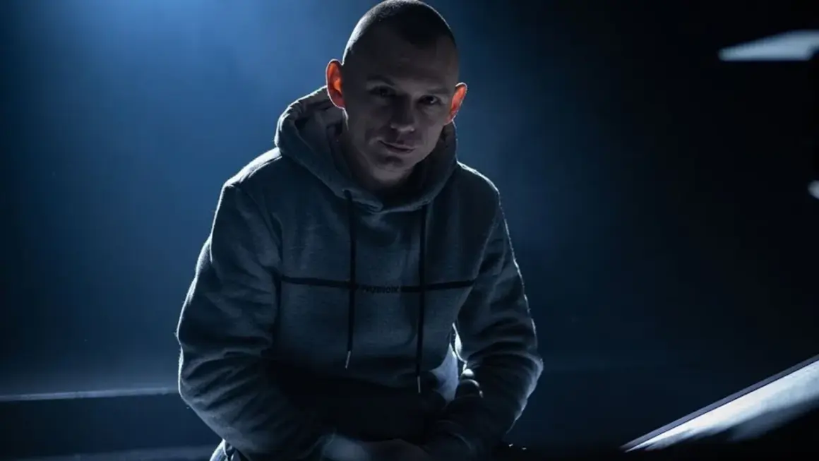 Mężczyzna w szarej bluzie z kapturem, oświetlony od góry, na ciemnym tle.