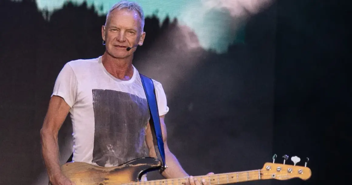 Sting gra na gitarze basowej w białej koszulce na tle sceny.