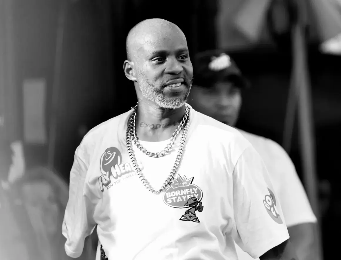 DMX w białej koszulce z łańcuchem na szyi, z tatuażem "EXODUS" na karku, uśmiecha się podczas występu.
