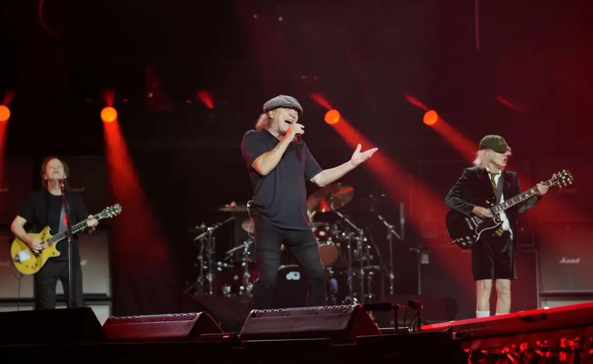 Zespół AC/DC na scenie, wokalista śpiewa, gitarzyści grają. Czerwone światła.