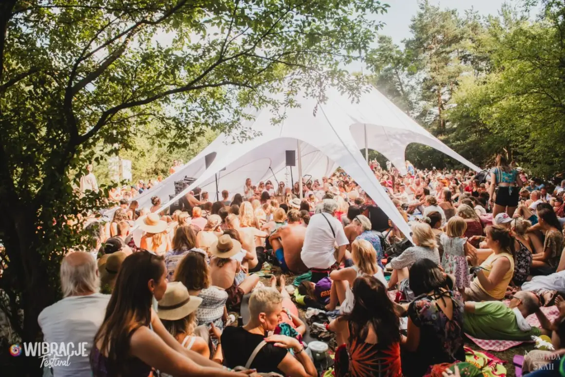 Tłum ludzi siedzi pod białym namiotem na festiwalu Wibracje, słuchając wystąpienia.