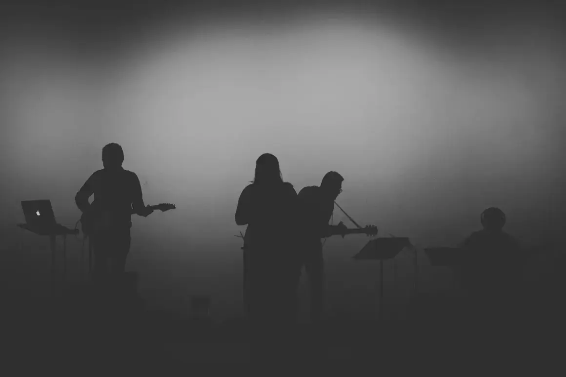 Sylwetki muzyków grających na gitarach i klawiszach na tle mglistego światła.