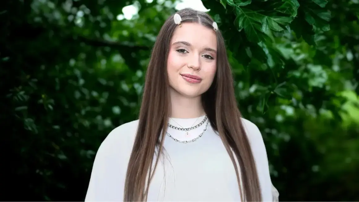 Uśmiechnięta Nicole Pniewska w białej bluzce, z długimi brązowymi włosami i delikatnymi spinkami, na tle zielonych liści.