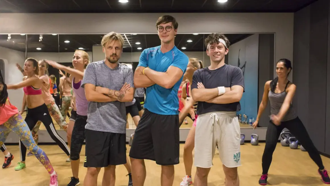 Trzech mężczyzn w strojach sportowych stoi z rękami skrzyżowanymi na piersi, w tle grupa kobiet ćwiczy.