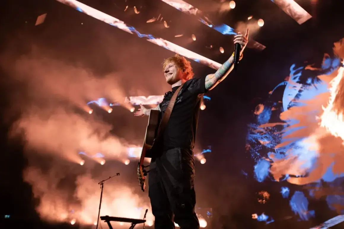Ed Sheeran na koncercie w Polsce, grając na gitarze i pozdrawiając publiczność.