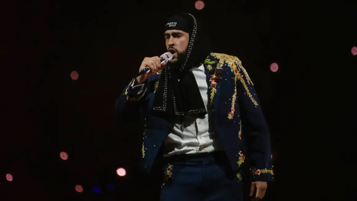 Bad Bunny w ozdobnej marynarce śpiewa na koncercie na ciemnym tle z rozmazanymi światłami.