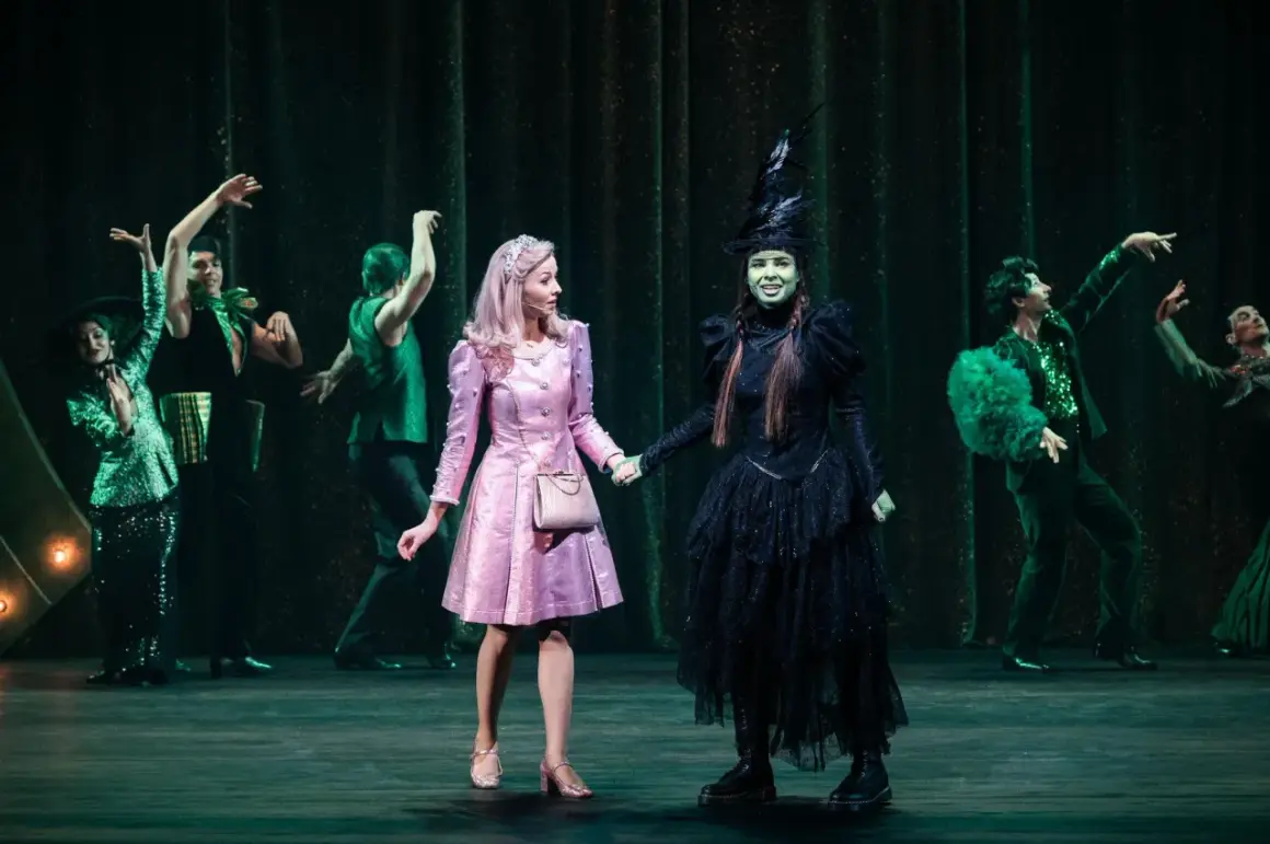 Elphaba w czarnej sukni i Glinda w różowej sukience trzymają się za ręce na scenie.