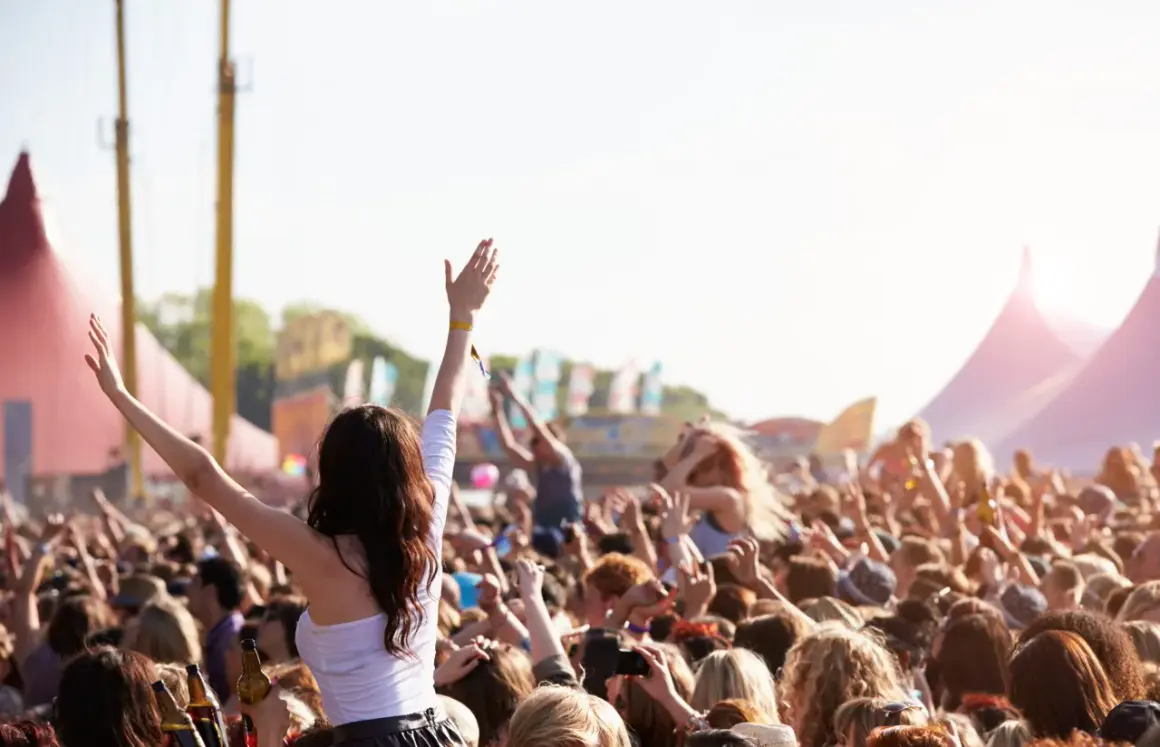 Tłum ludzi na festiwalu muzycznym, z rękami w górze, w tle namioty i sceny.