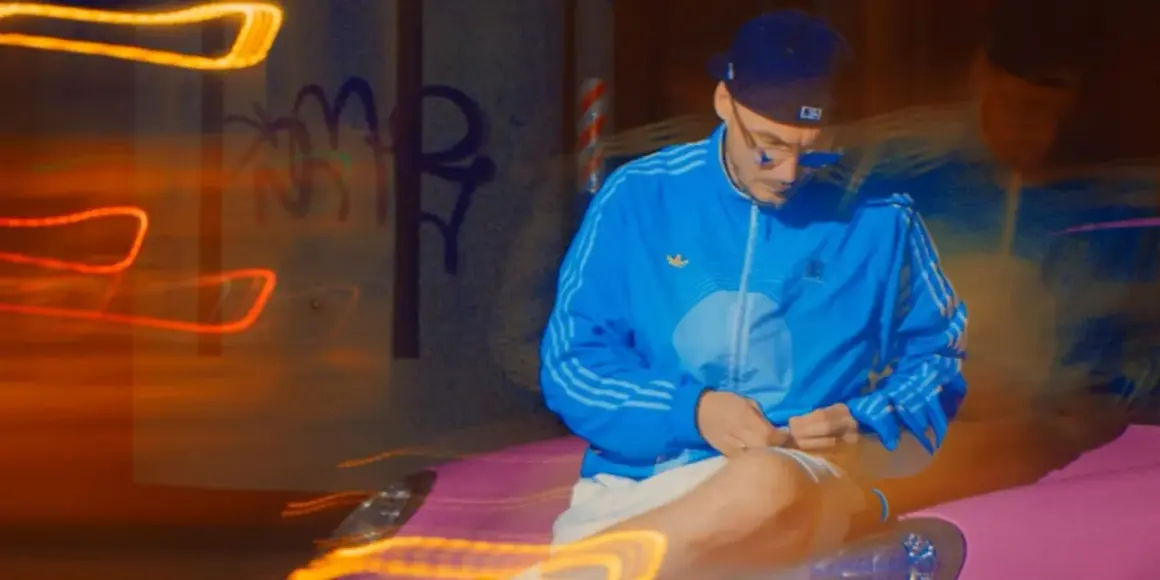 Mężczyzna w niebieskiej kurtce i czapce siedzi na tle nocnego miasta z neonowymi światłami.