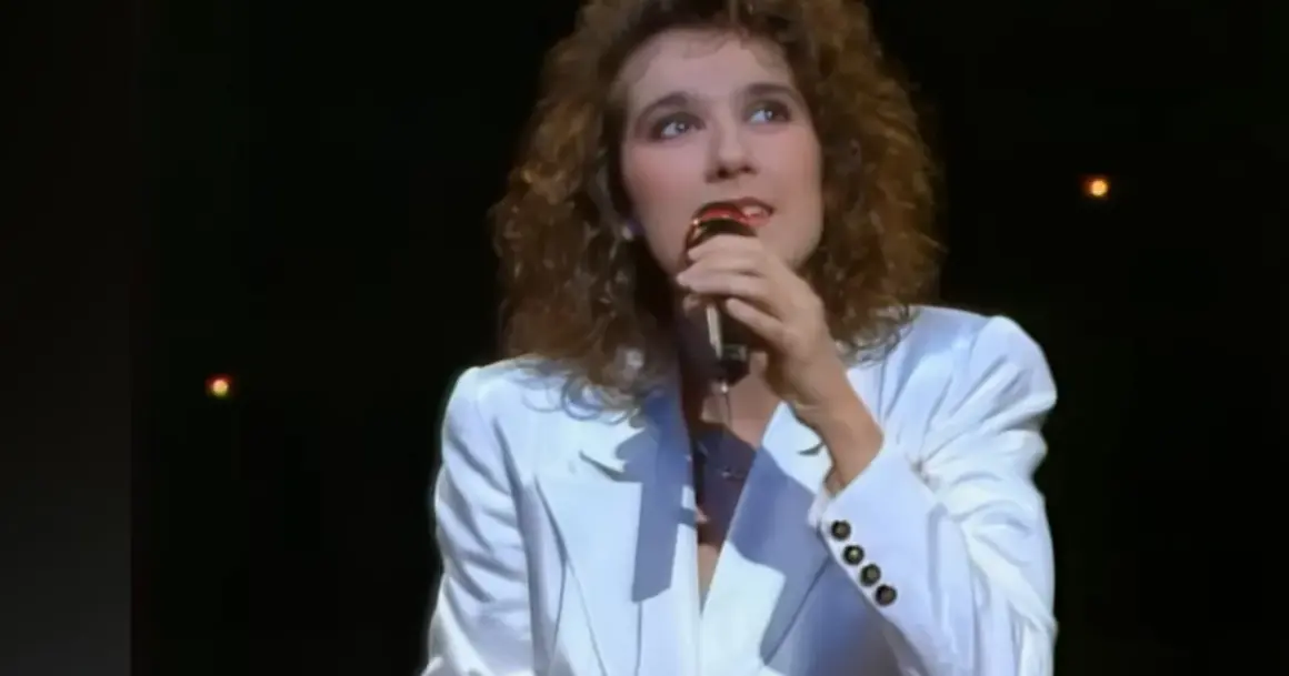 Celine Dion w białej marynarce śpiewa do mikrofonu na ciemnej scenie.