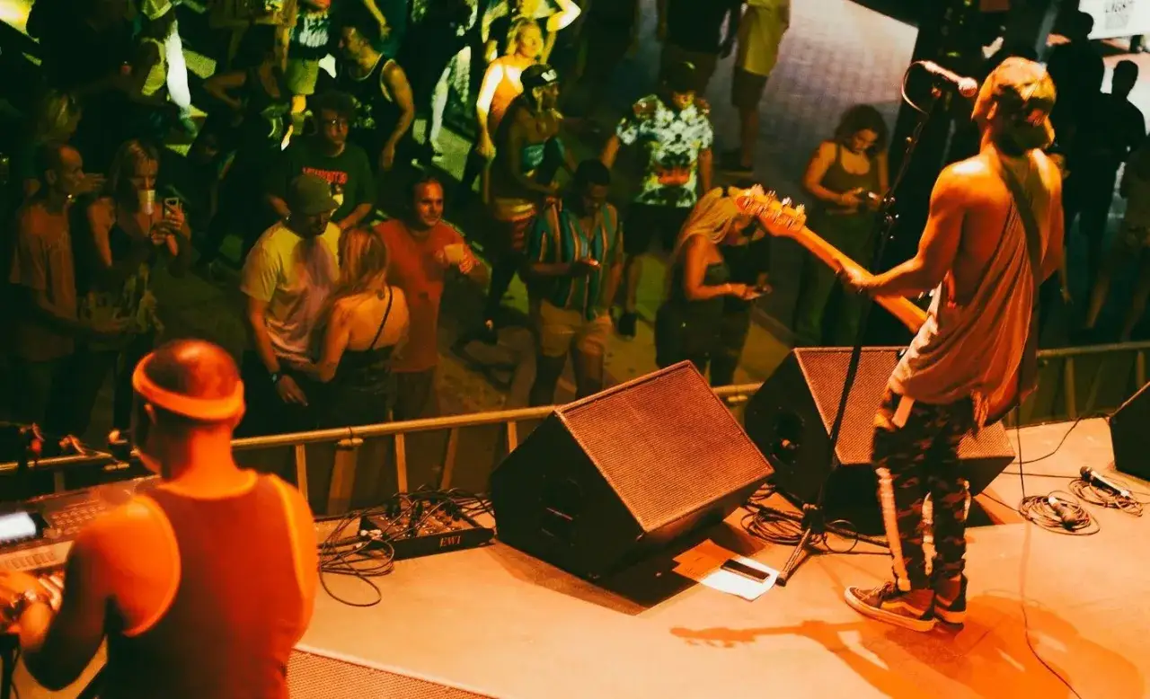 Koncert na żywo, muzyk gra na gitarze basowej, tłum bawi się w rytm muzyki. W tle widać sprzęt nagłośnieniowy, a na pierwszym planie DJ przy konsoli. To świetna ska zabawa!