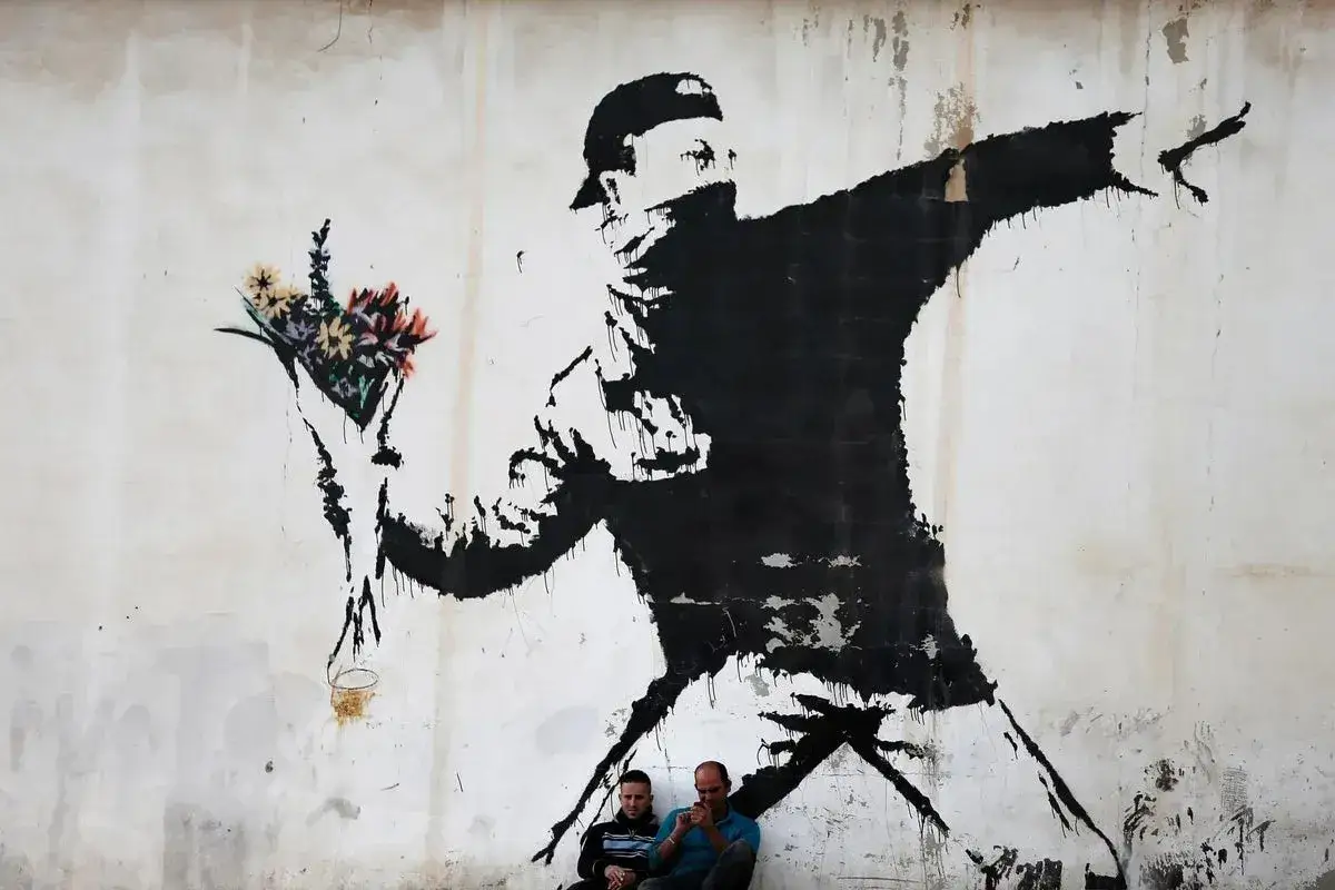 Banksy zdemaskowany: mural przedstawia zamaskowanego mężczyznę rzucającego bukiet kwiatów. Dwóch mężczyzn siedzi pod dziełem.